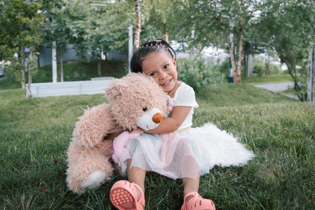 A smiling little girl hugging a brown teddy bear with some greenery in the background